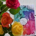 Anthology, 'You are Enough', with a cover of paint splashes, on white floor boards with yellow, red and orange roses in the foreground.